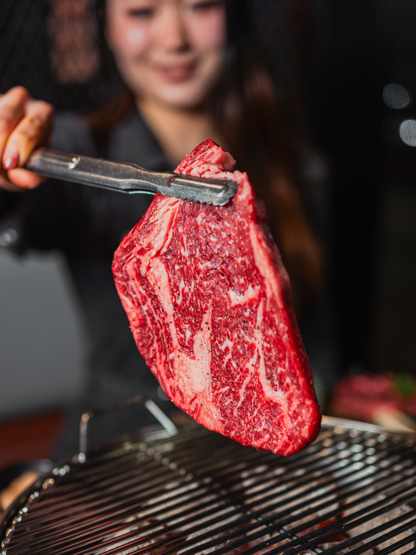 Server holding a thick marbled steak with tongs above the grill